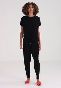 Zwart jumpsuit met korte mouwen, elastische taille en taps toelopende benen. Model draagt rode sandalen en grote ronde oorbellen.