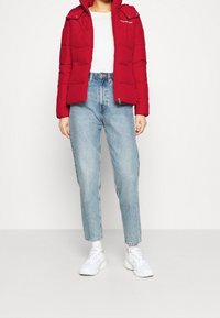 Rote Steppjacke mit Reißverschluss, Druckknopfkragen und Logodetail, kombiniert mit hellblauen Jeans mit geradem Bein und weißen Sneakers.