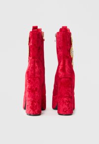 Bottes à cheville en velours rouge avec un large talon, présentant un accent doré près de la fermeture éclair et une surface texturée.