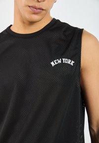 Schwarzes ärmelloses Mesh-Sportoberteil mit "NEW YORK" in weißen gestickten Buchstaben auf der oberen linken Seite. Texturiert und atmungsaktiv.