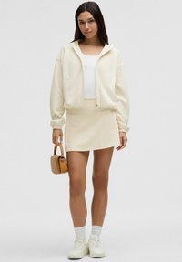 Hoodie cropped crema con cordini, top aderente bianco, gonna mini crema abbinata, sneakers bianche e piccola borsa color beige.