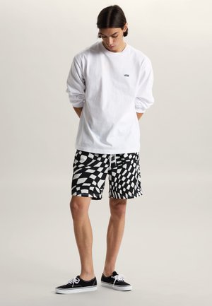 Mężczyzna w białej koszuli z długim rękawem, czarno-białych spodenkach z geometrycznym wzorem oraz czarnych trampkach, stojący z rękami za plecami, patrzący w dół.