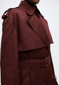 Bordeauxrode trenchcoat met een dubbelzijdig ontwerp, brede kraag, ceintuur in de taille en schouderepauletten, gemaakt van gladde stof.