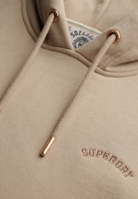 Beige hoodie met trekkoord, voorzien van gouden hardware accenten en geborduurd "SUPERDRY" logo. Zachte stof met een gladde textuur.