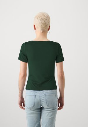 Personne aux cheveux blonds courts portant un haut vert foncé à manches courtes et un jean bleu clair, debout de dos devant un fond uni.