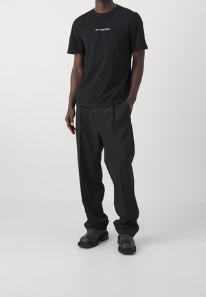 T-shirt noir en coton avec logo texte blanc, associé à un pantalon noir ample et des bottes noires épaisses, présentant une coupe décontractée et un design minimaliste.