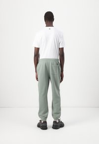 Witte katoenen T-shirt met logo op de achterkant, gecombineerd met groene sweatpants met een elastische tailleband en zijzak. Zwarte sneakers.