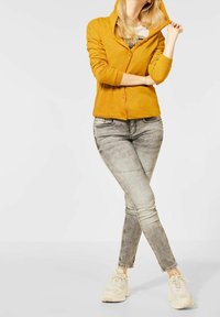 Cardigan à capuche jaune moutarde avec des boutons, associé à un jean skinny gris clair avec des accents cloutés et des baskets beiges.