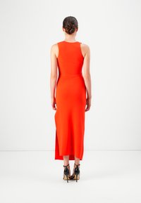 Frau mit dunklen Haaren im Dutt, die ein ärmelloses, figurbetontes rotes Kleid und schwarze High-Heels trägt, steht vor einem weißen Hintergrund.