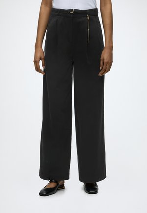 Person iført sorte, vidde bukser med høj talje og et tyndt bælte samt en hvid top, stående med armene afslappet ned langs siderne.