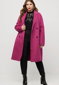 Cappotto lungo in misto lana fucsia con design doppiopetto, grandi bottoni neri e vestibilità ampia, indossato sopra un abbigliamento aderente nero e stivali.