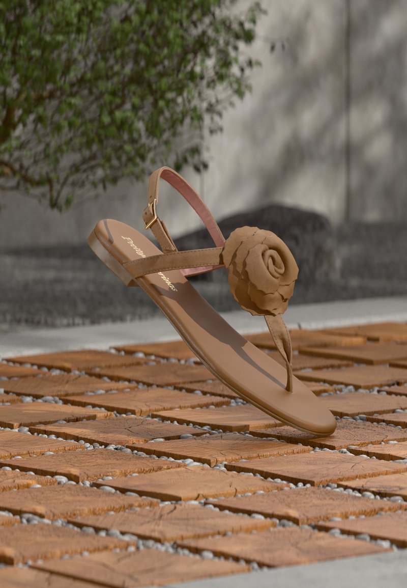 Καφέ επίπεδη σανδάλι με διακοσμητικό σε σχήμα τριαντάφυλλου, λουρί αστραγάλου και αγκράφα, τοποθετημένο σε ξύλινη επιφάνεια πλακιδίων με φυλλώδη φόντο.
