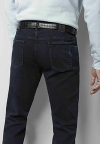 Donkerblauwe denim jeans met een slim fit ontwerp, voorzien van twee achterzakken en een gestructureerde zwarte riem. De stof lijkt glad en duurzaam.