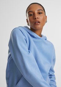 Svetlomodrá mikina z mäkkého materiálu, so predným vreckom, nastaviteľnou kapucňou so šnúrkou a voľným strihom s rebrovanými rukávmi.