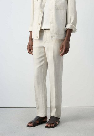 Person trägt hellbeige strukturierte Hose, passende Jacke mit Knöpfen und hochgekrempelten Ärmeln, weißes Top und schwarze offene Zehensandalen, steht auf grauem Boden.