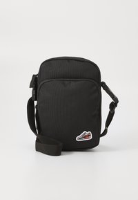 Svart crossover-veske laget av slitesterk stoff, med glidelås-lukking, justerbar stropp og en liten sneaker-patch på forsiden.