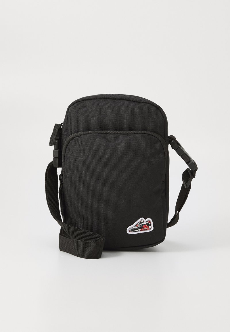 Črna crossbody torba iz trpežnega blaga, z zadrgo, prilagodljivim trakom in majhnim nalepko športne copate na sprednji strani.