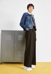 Jeansjacka med fårskinnskrage, grå topp, högmidjade bruna byxor och vita sneakers; står bredvid ett grått skåp på en gul golv.