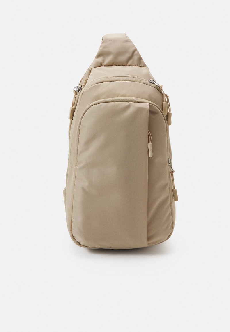 Sac bandoulière beige à une seule sangle avec plusieurs compartiments zippés et un dos rembourré, présenté sur fond blanc.