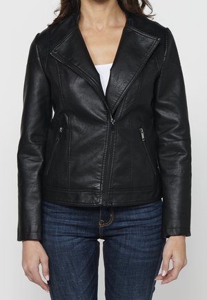 Femme portant une veste en cuir noire avec des poches zippées sur un haut blanc et un jean bleu, montrée du cou aux hanches.