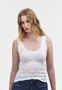 Vit ärmlös linne med rundad neck, tillverkat av lättviktig tyg. Kombinerat med blå denimjeans som har mässingsdetaljer.