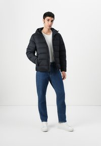Black puffer jacket with a hood, featuring quilted panels. Paired with blue denim jeans and white sneakers. Plain white t-shirt underneath.
