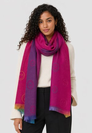Woman with curly hair wearing cream sweater, black pants, and long pink and purple scarf with smiley face patterns and multicolor fringes.