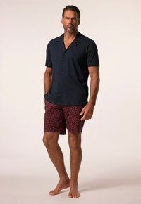 Marinblå skjorta med korta ärmar; kombinerad med röda shorts med ett litet kontrasterande blommönster. Modell står barfota.