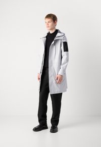 Jachetă impermeabilă de culoare gri deschis, cu glugă, două buzunare frontale și un petic negru pe mânecă, purtată cu pantaloni și încălțăminte negri.