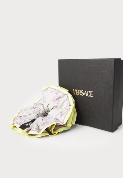Versace scrunchie v svetlo vijolični barvi z igrivim vzorcem, obrobljena z rumeno, ob črni teksturirani škatli z vtisnjenim logotipom.