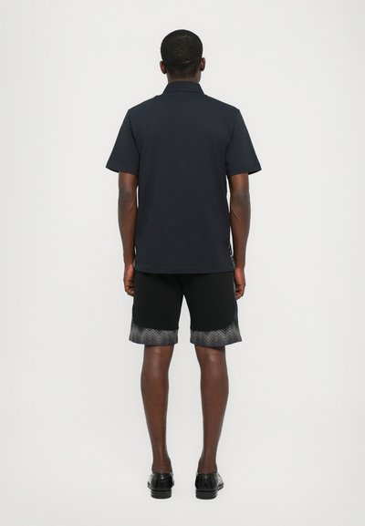 Tricou polo cu guler bleumarin, asortat cu pantaloni scurți negri cu un model zigzag la margine. Purtați cu pantofi negri. Vedere din spate.