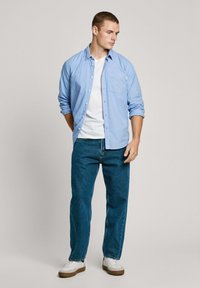 Ljusblå skjorta med knappar, vit T-shirt under, blå jeans och vita sneakers. Skjortan har en enda bröstficka.