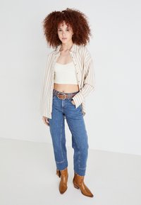 Camicia oversize a righe beige e bianche sopra un top corto bianco, abbinata a jeans a vita alta blu e stivaletti marroni.
