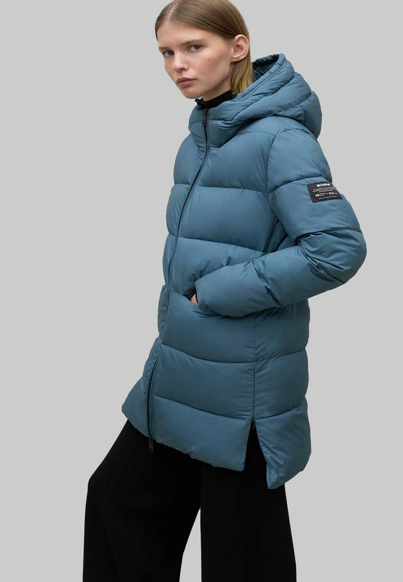 Chaqueta acolchada color azul verdoso con capucha, que presenta secciones acolchadas, bolsillos laterales y cierre de cremallera. Incluye una etiqueta de tela negra en la manga.