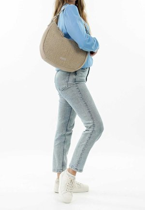 Kobieta w jasnoniebieskiej bluzce i dżinsach, nosząca białe trampki, trzymająca beżową, teksturowaną torbę na ramię.