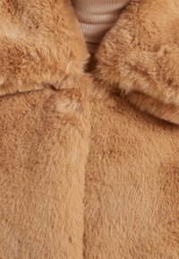 Faux-Fur-Mantel in einem sanften Goldbraun, mit flauschiger Textur und rundem Kragen. Der Stoff zeigt einen dezenten Glanz mit dichten, kurzen Fasern.