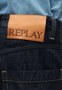 Σκούρο μπλε τζιν από τζιν ύφασμα με ραμμένη καφέ δερμάτινη ετικέτα που φέρει την επωνυμία "REPLAY" στη μέση.