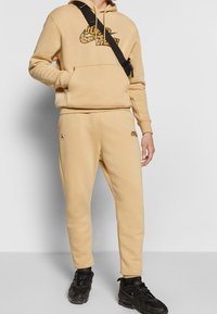 Beiger Hoodie mit schwarzem Nike-Logo im Leopardenmuster, kombiniert mit beigen Jogginghosen mit schwarzen Akzenten. Dazu schwarze Sportschuhe.