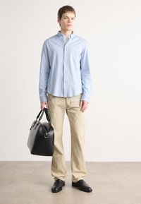 Lichtblauwe button-up shirt, beige broek, zwarte leren tas en zwarte instap schoenen, staande op een neutrale achtergrond.