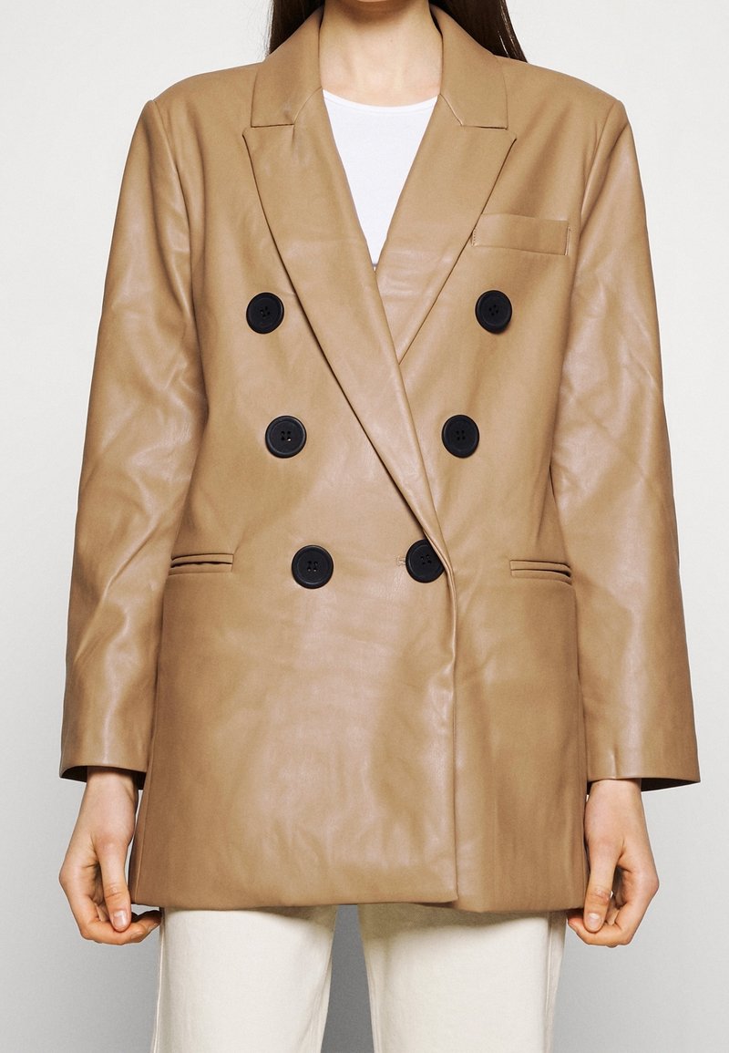 Femme portant un blazer en cuir marron clair à double boutonnage avec des boutons noirs, une chemise blanche en dessous et un pantalon de couleur claire.
