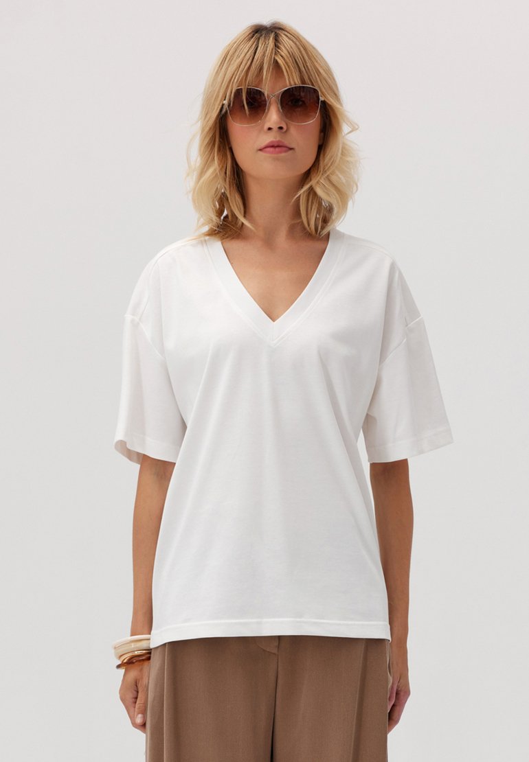 Biała bawełniana koszulka z dekoltem w serek, z krótkimi rękawami, luźnym krojem i prostym dołem. Noszona z beżowymi spodniami w wysokim stanie i zwielokrotnionymi bransoletkami.