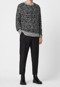 Person i grå och svart djurmönstrad tröja, svarta cropped byxor och glänsande svarta snörkängor mot vit bakgrund.