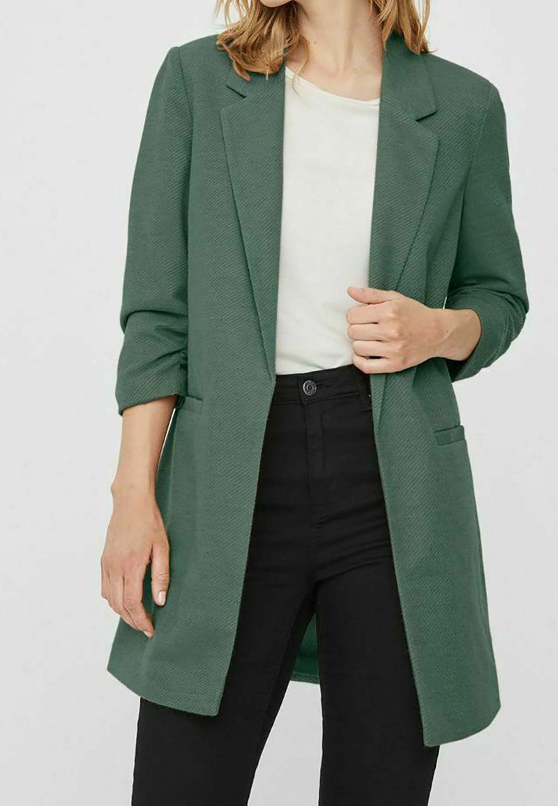 Groene, gestructureerde blazer met een opstaande kraag en opgerolde mouwen, voorzien van een enkele voorkleurige zak, gecombineerd met een witte shirt en zwarte broek.