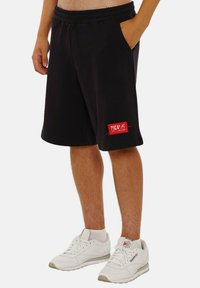 Schwarze Shorts mit elastischem Bund, Seitentaschen und einem roten Logo-Patch. Getragen mit weißen Turnschuhen mit grauen Akzenten.