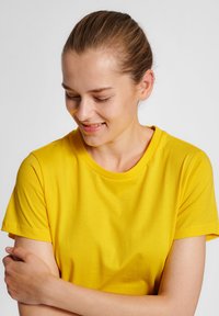 Kortärmad gul bomullströja med rundad halsringning, slät textur och figurnära silhuett, bärs av en person med korslagda armar.