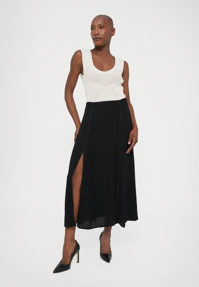 Fustă maxi neagră cu o tăietură pe lateral, asortată cu un top din tricou crem cu textură și pliuri. Toate acestea sunt completate de niște tocuri stiletto.