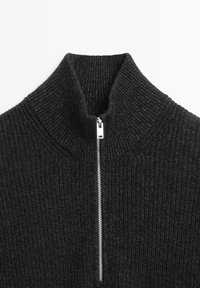 Zwarte geribbelde trui met hoge kraag en halve ritssluiting. De stof heeft een gestructureerd gebreid patroon. Voorop bevindt zich een zilveren rits.