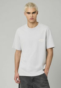 T-shirt en coton gris clair avec un col ras du cou, des manches courtes et le texte "Ongoing Project" dans une couleur d'accent subtil sur la poitrine.