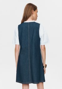 Denim jurk, donkerblauw, A-lijn vorm, mouwloos, met zichtbare stiksels, gedragen over een wit T-shirt, achterkant zichtbaar.