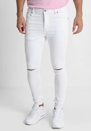 Mann trägt weiße Slim-Fit-Jeans mit Rissen, weiße Sneaker und ein pinkes Hemd und steht vor einem weißen, schlichten Hintergrund.